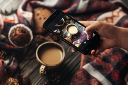 hand holding phone taking photo of stylish winter flat lay coffee cookies and spices on wooden rustic background. cozy mood autumn. instagram blogging workshop concept.