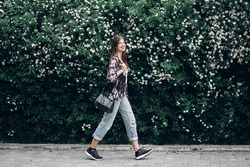 happy hipster woman with black bag walking on background of blooming bush with white flowers of spirea. boho girl smiling in floral clothes. space for text. spring summer time