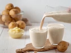 Potato milk pouring into glass on white wooden background. Pouring vegan milk in glass, with potato puree and potato tubers on background. Copy space. Home made potato milk made from boiled potatoes