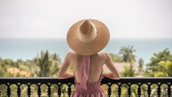 Back view of woman in pink dress and straw hat standing on a hotel balcony, ocean view.