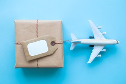 Flat lay of wrapped package with blank tag and airplane model on pastel blue background. Shipping logistics transport minimal creative concept.