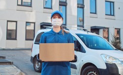 Portrait of Caucasian young female delivery postal worker in gloves and medical mask stepping in front of camera with parcel in hands. Close up of pretty happy woman handing carton box to cam outdoor.