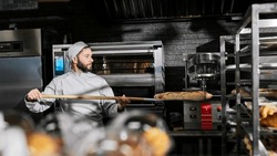 Caucasian male baker in apron and hat taking out just-baked baguettes from oven and putting on shelf in kitchen of bakehouse. Man working in bakery and baking fresh bread. Cooking profession.