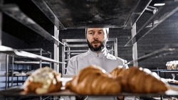 Portrait shot of Caucasian handsome man in hat and uniform working in bakehouse and checking croissants. Male baker with beard making fresh pastries in kitchen of bakeshop in morning.