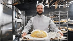 Portrait of handsome Caucasian male in white apron and hat standing in front of camera, holding tray with fresh just-baked bread and smiling in kitchen of bakehouse. Man in bakeshop showing baking.
