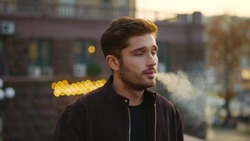 Portrait of handsome guy blowing smoke from e-cigarette outdoors. Closeup hipster man smoking electronic cigarette in urban background. Attractive male person vaping outside.