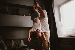Dad holds the baby in the air by the legs, funny home photos. Authentic lifestyle and brown tinting