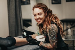 Charming girl with dreadlocks and tattoos stuffs a tattoo on a woman's leg in a tattoo parlor, creative small business and tattoo master. Hipster woman in gloves draws a tattoo