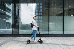 Side view of young smiling businesswoman with cycling helmet on her head driving an electrical push scooter
