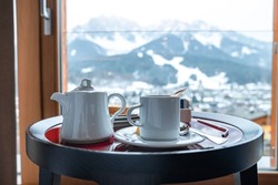 Close-up of white tea cup and kettle on stool. Breakfast served with ceramics on table at resort. Mountains seen from window in background during winter.