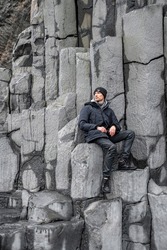 Tourist sitting on beautiful basalt columns. Thoughtful man exploring rocky formation on seashore. Scenic view of gray patterned stones at famous Reynisfjara Beach.