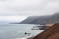 High angle view of steep brown slope. Scenic view of sea waves rushing at black sand beach against cloudy sky. Beautiful scenery of volcanic valley during foggy weather.