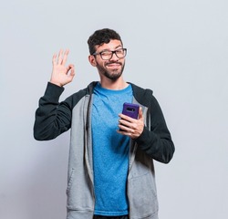 Pleasant person holding cellphone and winking, smiling boy with cellphone making ok sign, concept of positive person with cellphone closing fingers in ok sign