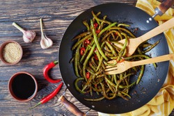 Spicy Chinese Sichuan stir-fried Green Beans in a wok with spatulas, on a wooden table with kitchen towel and ingredients, view from above, flat lay