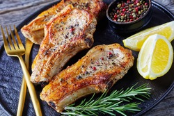 close-up of broiled pork rib chops with lemon wedges, rosemary, peppercorns and golden fork and knife on a black platter on a rustic wooden table, horizontal view