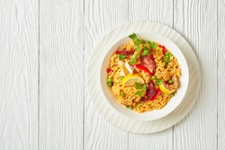 portion of spanish chicken paella with valencian bomba rice, chicken thigh meat, chorizo sausages, vegetables and spices served on a white plate on a white wooden table, flat lay