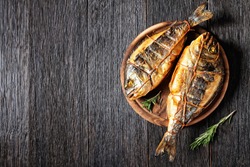 hot smoked gilt-head sea bream, orata fish on a wooden round cutting board on a dark wooden table, horizontal view from above, flat lay, free space