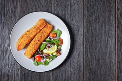 Panko crusted baked salmon fillets with spinach tomato cucumber olives salad on a plate on a dark wooden table with fork and knife,  view from above,  flat lay, free space