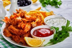breaded and deep fried prawn tails with potato fries, tomato sauce and tartar sauce on a white plate, horizontal view, close-up