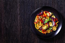 roasted red, orange and yellow pepper slices with red onion wedges and zucchini on black plate on dark oak wood table, horizontal view from above, flat lay, free space