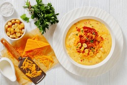 savory creamy pumpkin soup topped with fried bacon, shredded cheddar cheese and salty croutons in white bowl on wood table with ingredients, horizontal view from above, flat lay