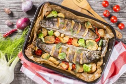 delicious trout fish baked with potatoes, broccoli, lemon, tomatoes and spices in baking dish on a wooden background, top view