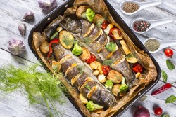 delicious trout fishes baked with potatoes, broccoli, lemon, tomatoes and spices in baking dish on a wooden background, view from above