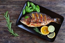 delicious roasted dorado or sea bream fish with lemon and orange slices, spices, fresh rosemary and spinach on black plate on old dark wooden table view from above