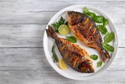 delicious grilled dorado or sea bream fish with lemon and orange slices, spices, fresh parsley and spinach on white platter on old wooden table, horizontal view from above