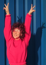 Happy young woman with curly hair in magenta suit and sunglasses posing on blue background.