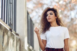 Beautiful young arabic woman with black curly hairstyle. Arab girl in casual clothes in the street. Happy female wearing white t-shirt and checked pants.