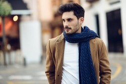 Young man wearing winter clothes in the street. Young bearded guy with modern hairstyle with coat, scarf, blue jeans and t-shirt.