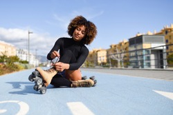 Young smiling black girl sitting on bike line and puts on skates. Woman with afro hairstyle rollerblading on sunny day