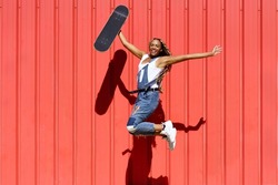 Black woman dressed casual, wtih a skateboard jumping with happiness on red urban wall background.