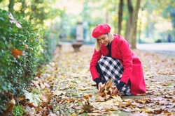 Positive female in checkered skirt and red outerwear picking fallen leaves from path in autumn park