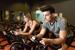Attractive woman and man biking in the gym, exercising legs doing cardio workout cycling bikes. Couple in a spinning class wearing sportswear.