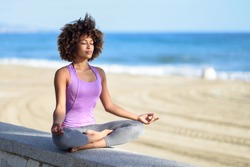 Black woman, afro hairstyle, doing yoga asana in the beach with eyes closed. Young Female wearing sport clothes in lotus pose with defocused background.