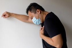Side View Of An Sick Asian Man In A Medical Disposable Mask, Feeling Difficulty Breathing, Standing Against Wall