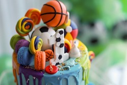 Creative birthday cake with colourful frosting decorated with mini basketball, football, macarons and lollipop sweet candy. Soft focus image, high angle view.