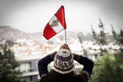 woman with chullo in the head is holding peru flag raising both hands on park background
