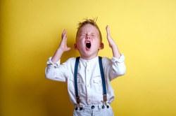 Child shouting loud. Stylish little child boy holding hands near head. Portrait of shocked, angry and emotional little boy  isolated on yellow background.