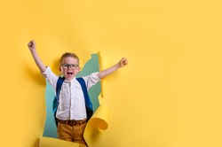 Child boy with book and bag breaking through yellow paper wall. Happy 
smiling kid go back to school, kindergarten. Success, motivation, winner, genius concept. Little kid dreaming to be superhero