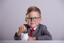 Child boy in suit and glasses putting coin into piggy bank