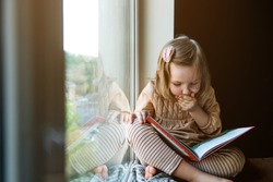Little girl is reading a book while sitting near window