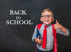 Happy smiling boy in uniform with book and bag. Surprised kid in glasses go back to school. Success, motivation, winner, genius concept. Businessman against blackboard background