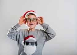 Merry christmas and happy new year. Happy smiling kid in a red hat and glasses. School child opens his eyes in surprise. Stylish boy in modern sweater with Santa Clause face. Funny kid portrait.