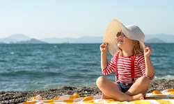 Funny little girl in a big striped hat on the beach. Kid in a hat and sunglasses on the beach. Happy little child on summer vacation.
