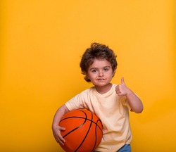 Portrait of a little boy with basketball isolated on yellow background. Small kid basketball player with thumb up. Sport child playing game. Kid activities. Little basketballer. Sports equipment