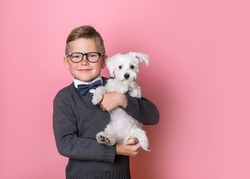 Child in bow tie, shirt and glasses with funny little white dog in hands. Cute friends lovingly embraced. pet dog and school kid isolated on pink background.