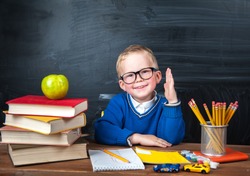 Happy cute clever boy is sitting at a desk in a glasses with raising hand. Child is ready to answer with a blackboard on a background. First time to school. Back to school. Apple and books on desk
